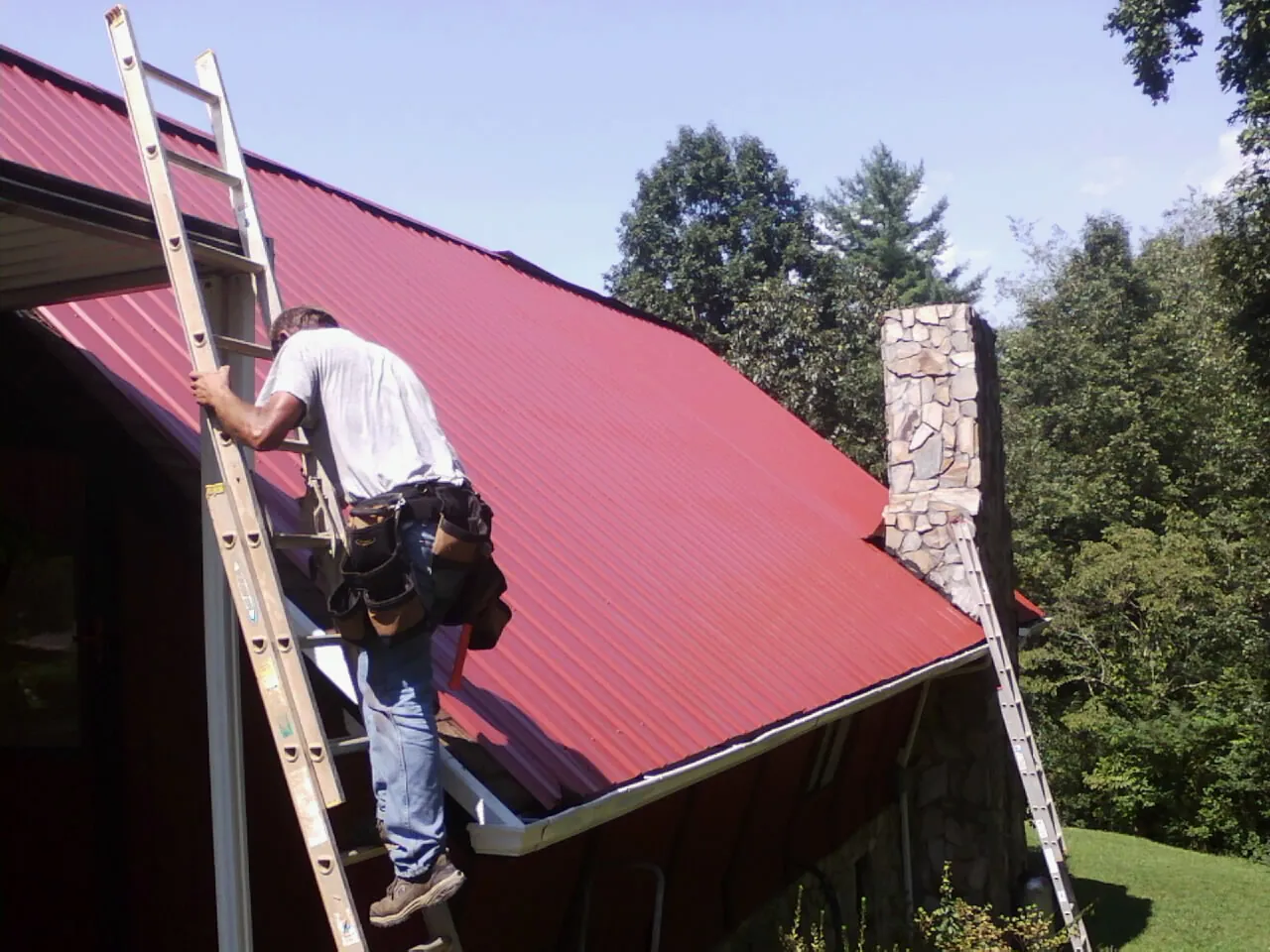 Roofer on a ladder beside a red metal roof and stone chimney — Soffit & Fascia Repair by Roofing Brothers in Humarock